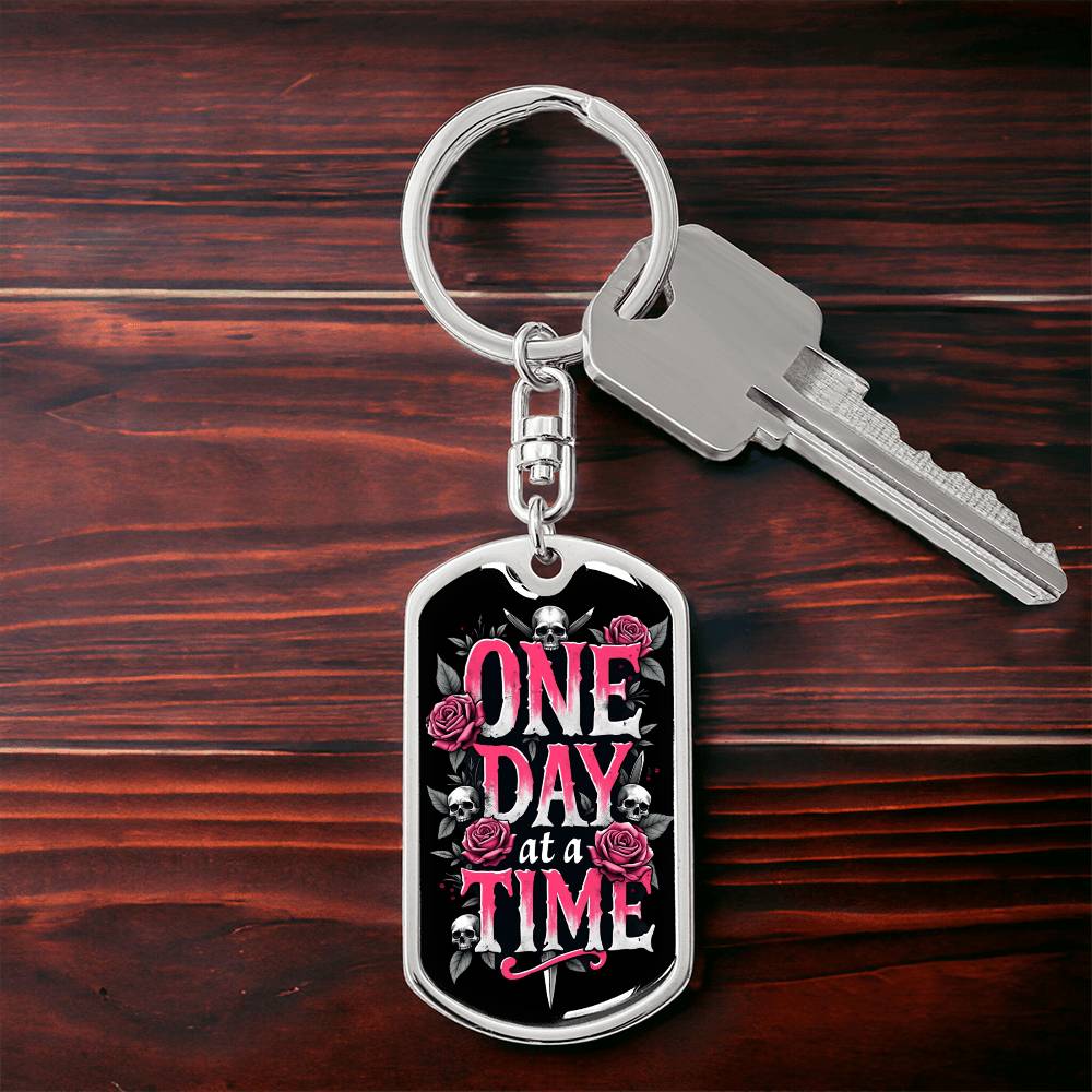Silver dog tag keychain with black graphic and pink text, on keyring