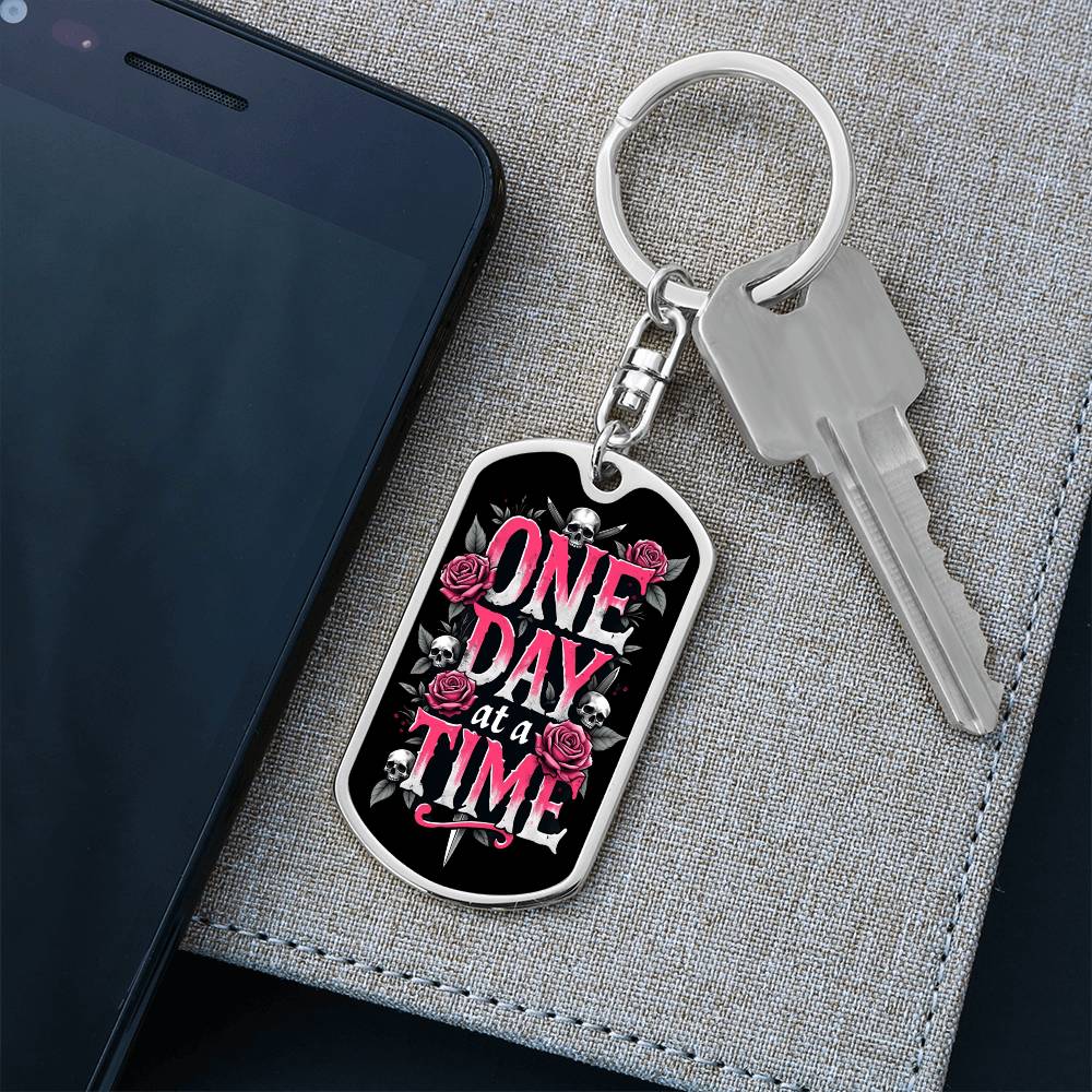 Silver dog tag keychain with black graphic and pink text, on split keyring