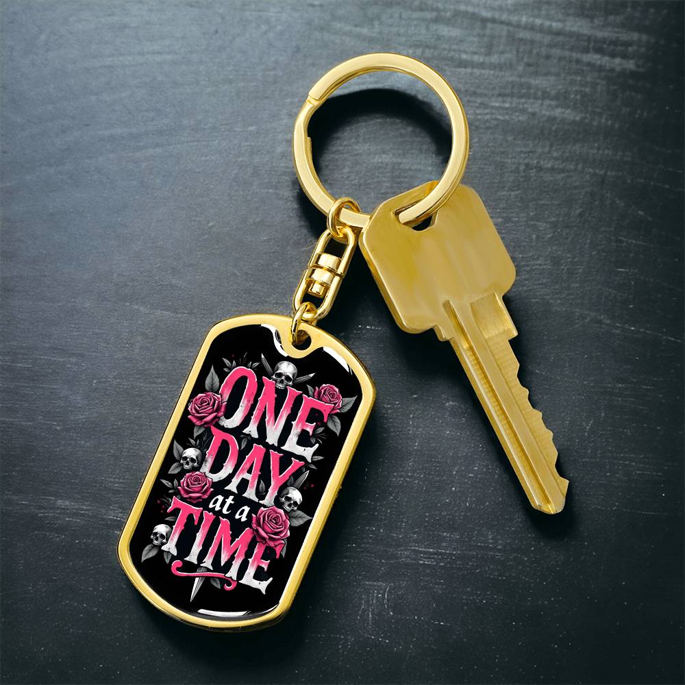 Gold dog tag keychain with black graphic and pink text, with key attached