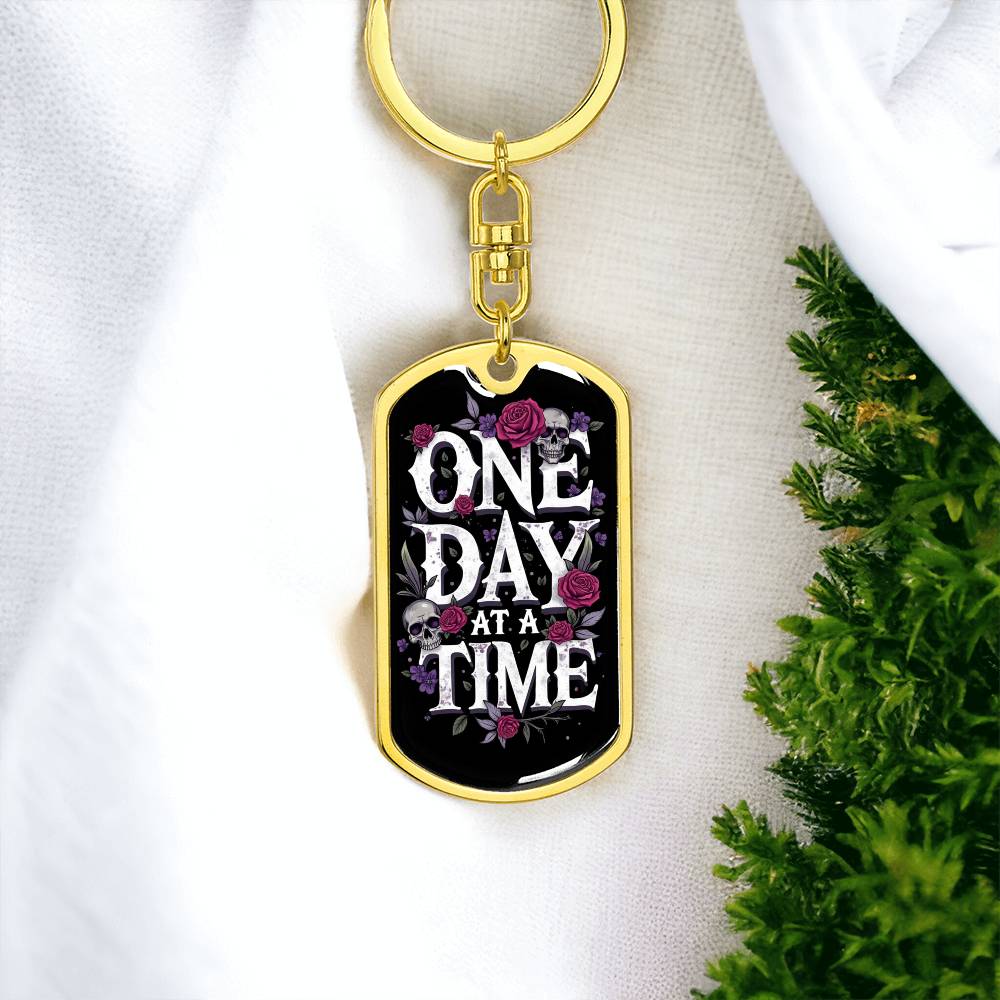 Gold dog tag keychain with black enamel face, floral skull design and “ONE DAY AT A TIME”