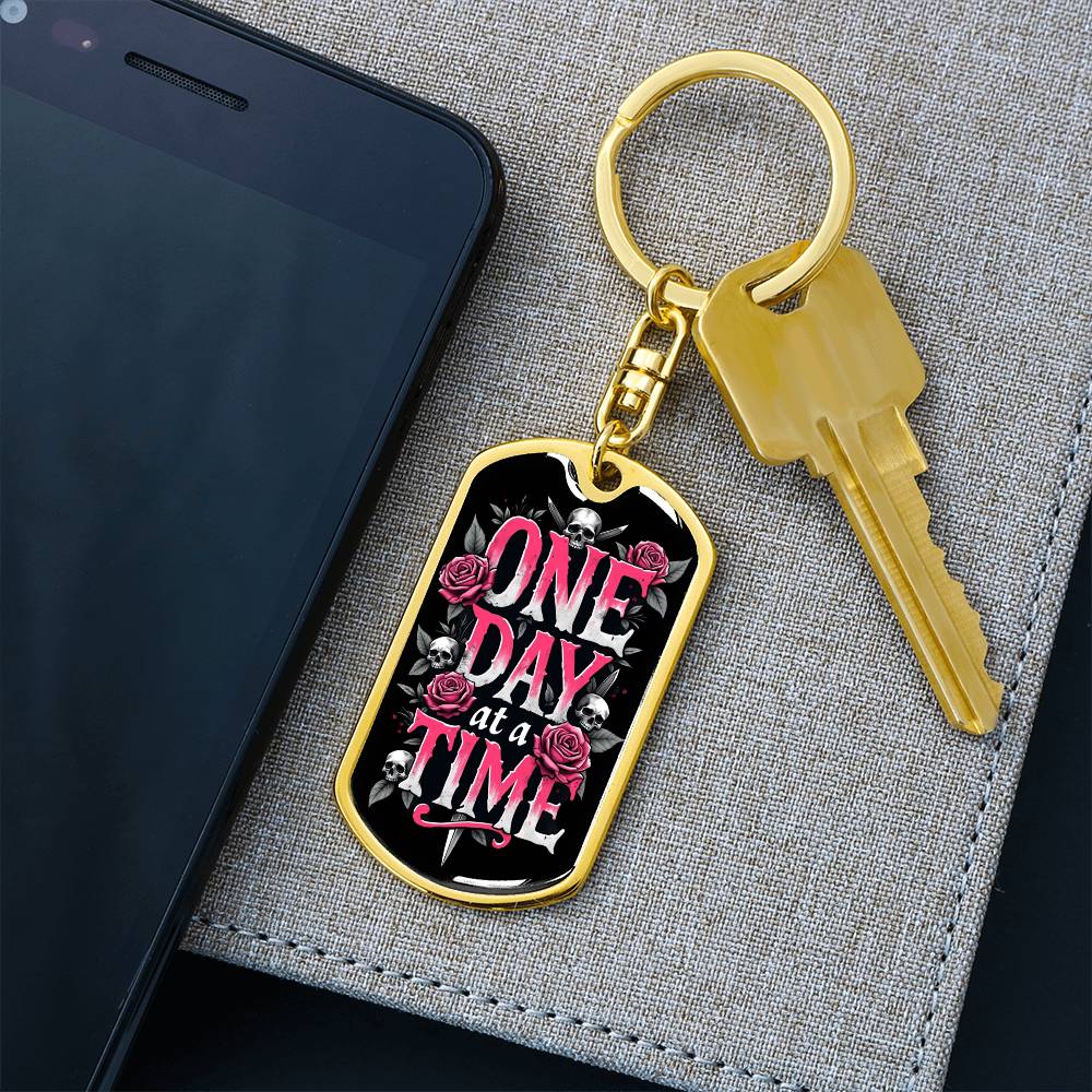 Gold dog tag keychain with black graphic and pink text, with key attached