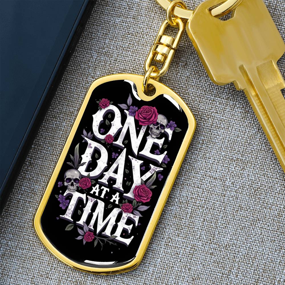Gold dog tag keychain with “One Day At A Time” front and attached house key