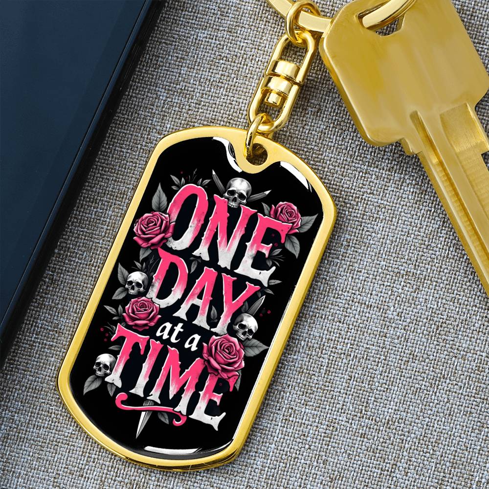 Gold dog tag keychain close-up with black face showing One Day at a Time, roses, skulls