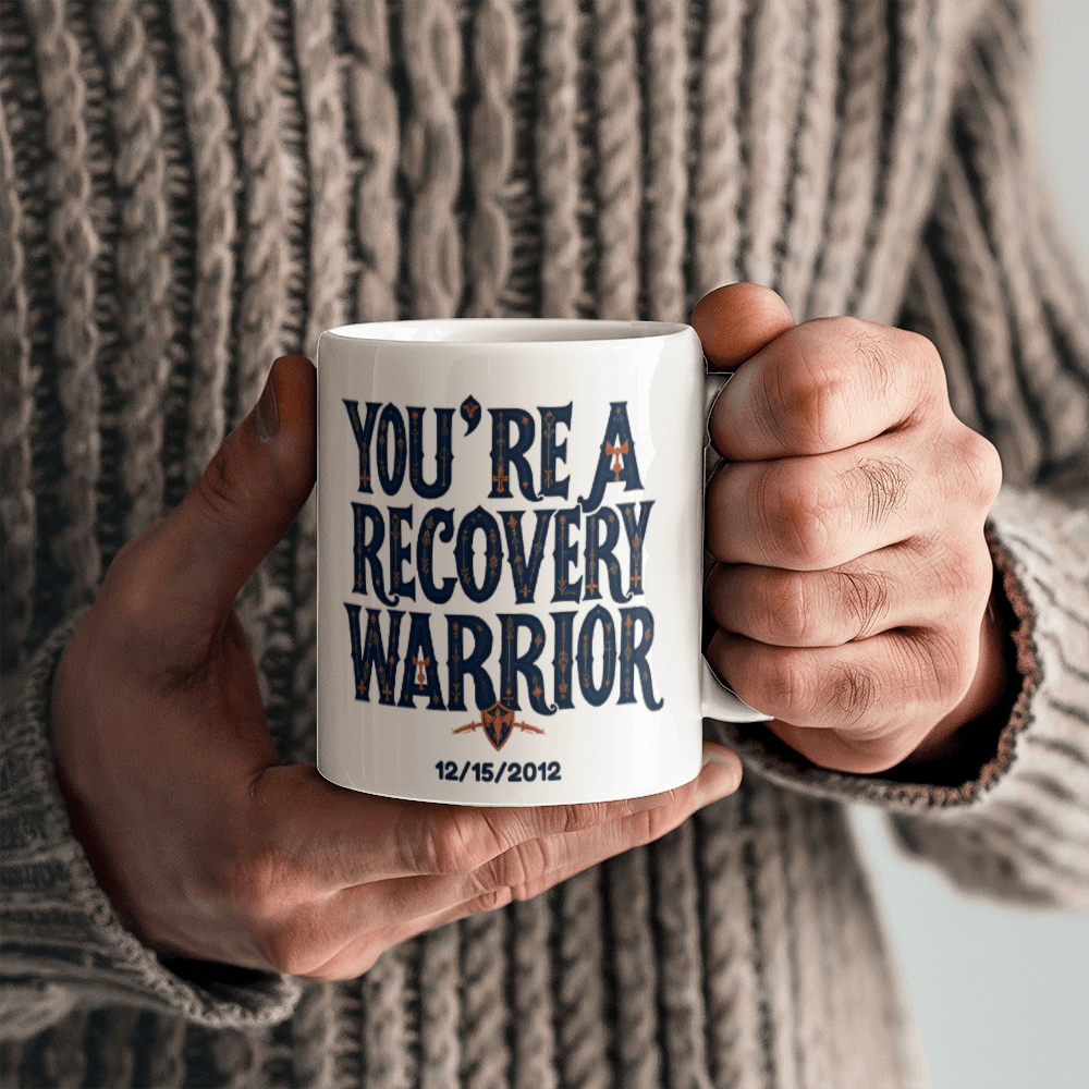 White ceramic coffee mug with “You’re a Recovery Warrior” graphic, held outdoors