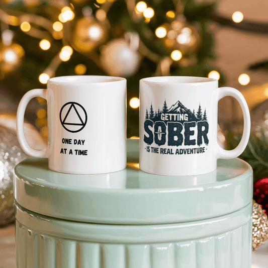 Two white ceramic mugs: 'ONE DAY AT A TIME' and 'GETTING SOBER' with pine trees