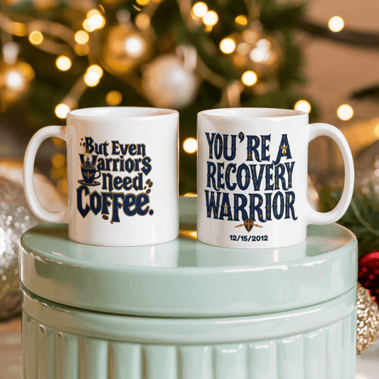 Two white coffee mugs on a stand; one reads “You’re a Recovery Warrior” with date